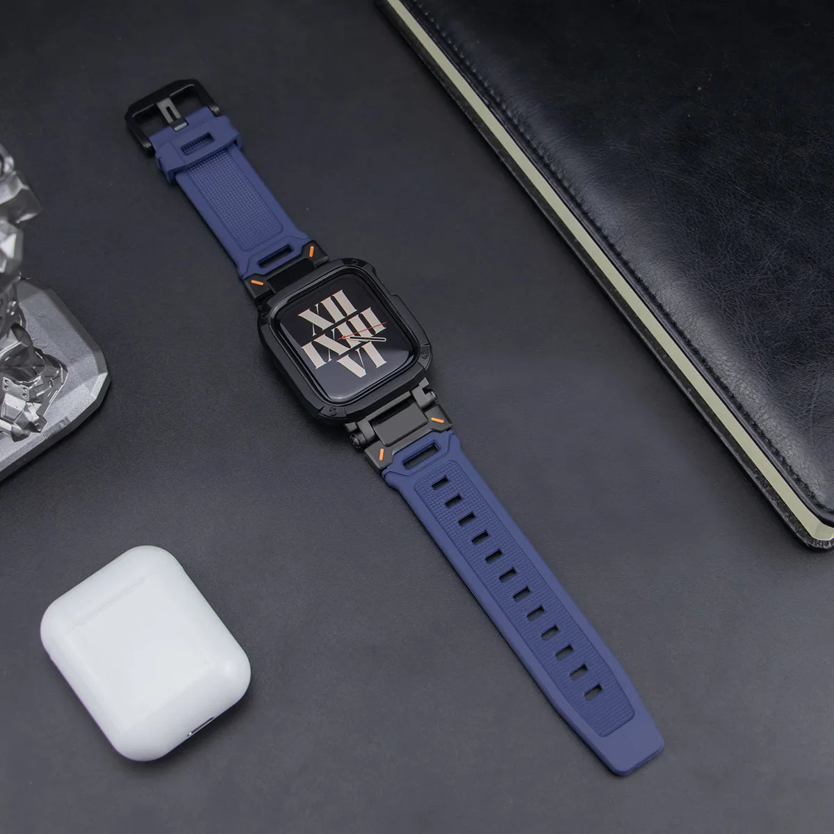navy blue apple watch band and black case, laid down on a black table next to air pods and a black notebook