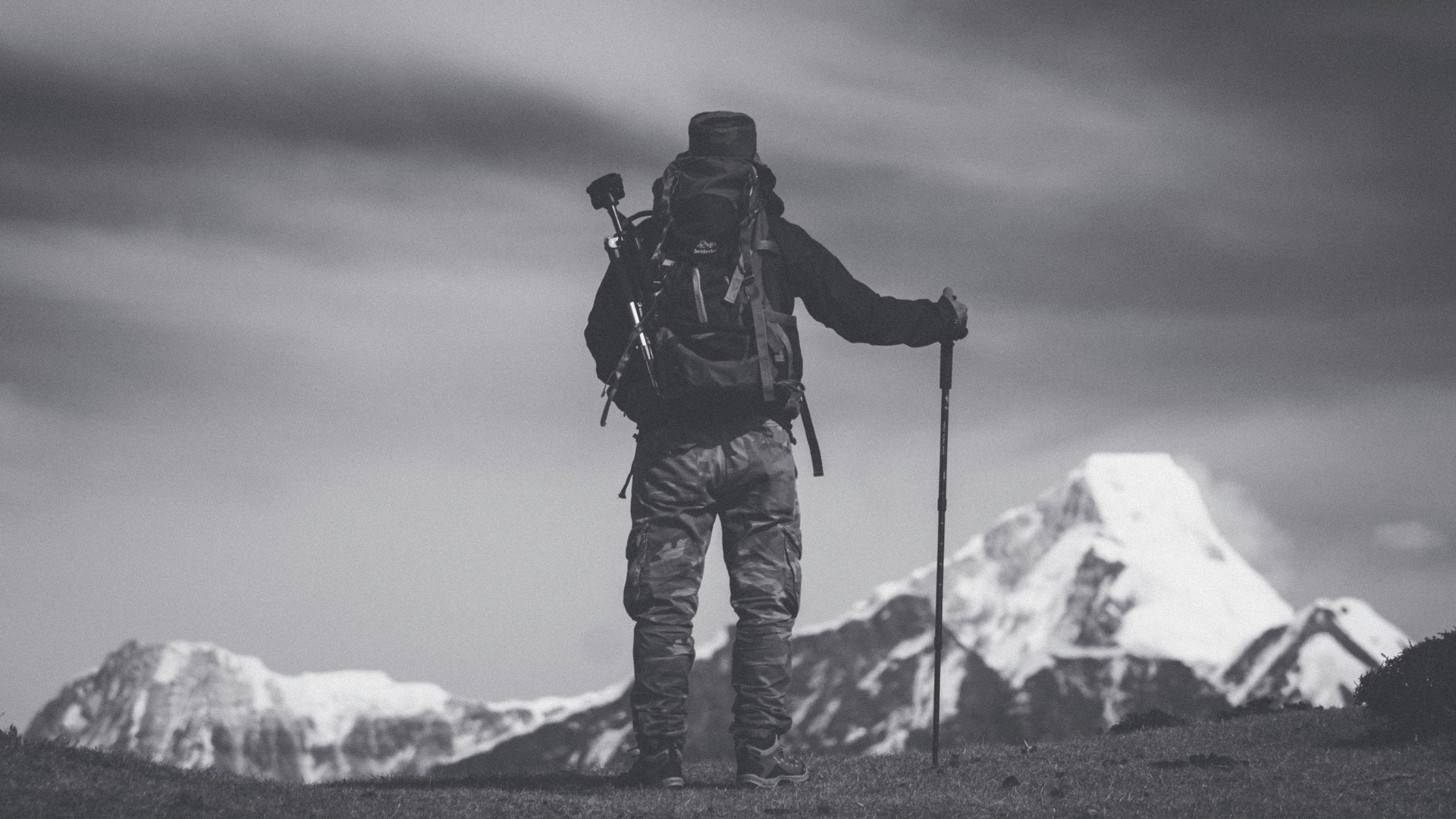 Person with a backpack and hiking poles standing in front of snow-capped mountains