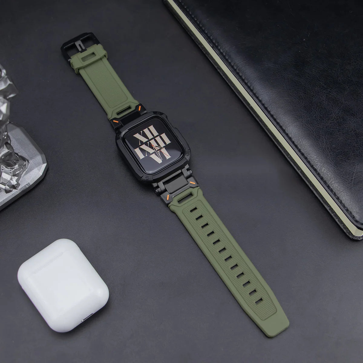 army green apple watch band and black case, laid down on a black table next to air pods and a black notebook