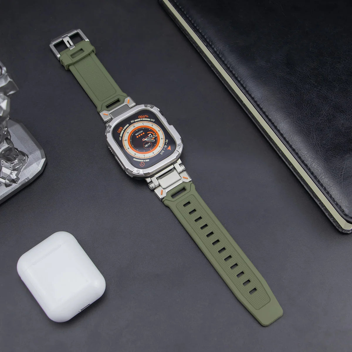 army green apple watch band and grey rugged case, laid down on a black table next to air pods and a black notebook