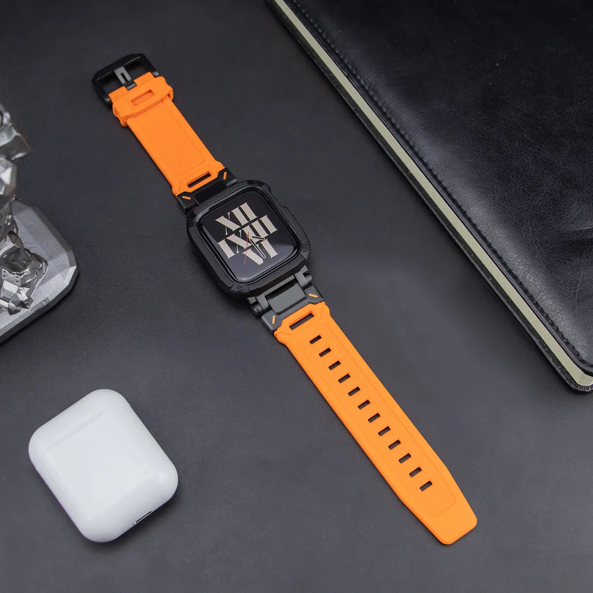orange apple watch band and black case, laid down on a black table next to air pods and a black notebook