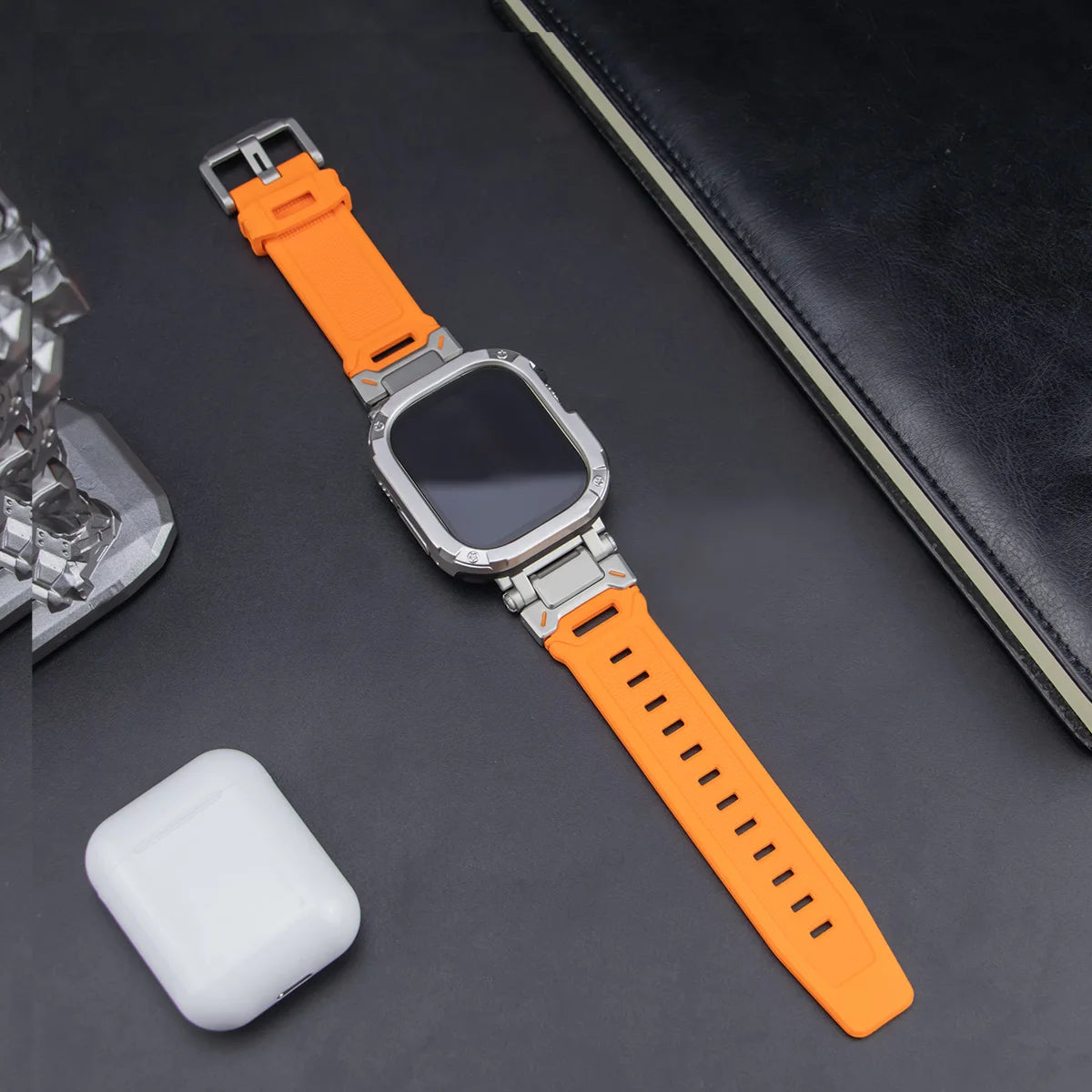 orange apple watch band and grey rugged case, laid down on a black table next to air pods and a black notebook