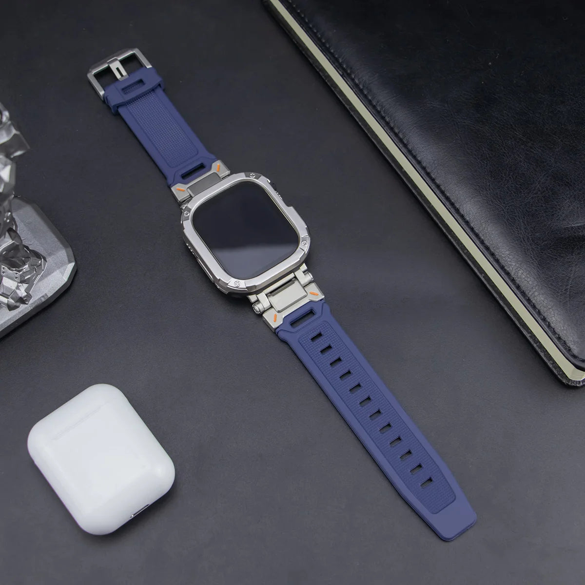navy blue apple watch band and grey case, laid down on a black table next to air pods and a black notebook