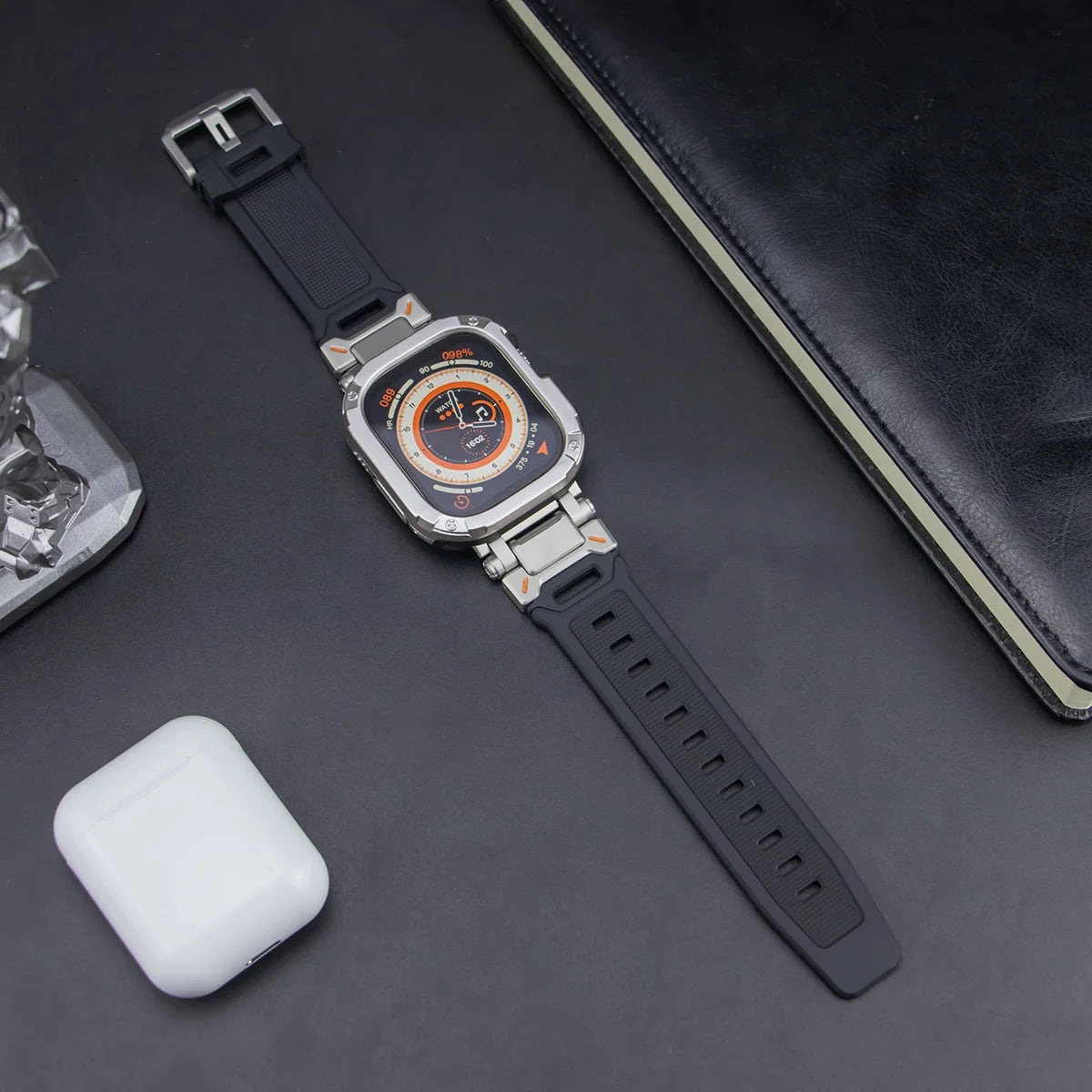 black apple watch band and grey rugged case, laid down on a black table next to air pods and a black notebook